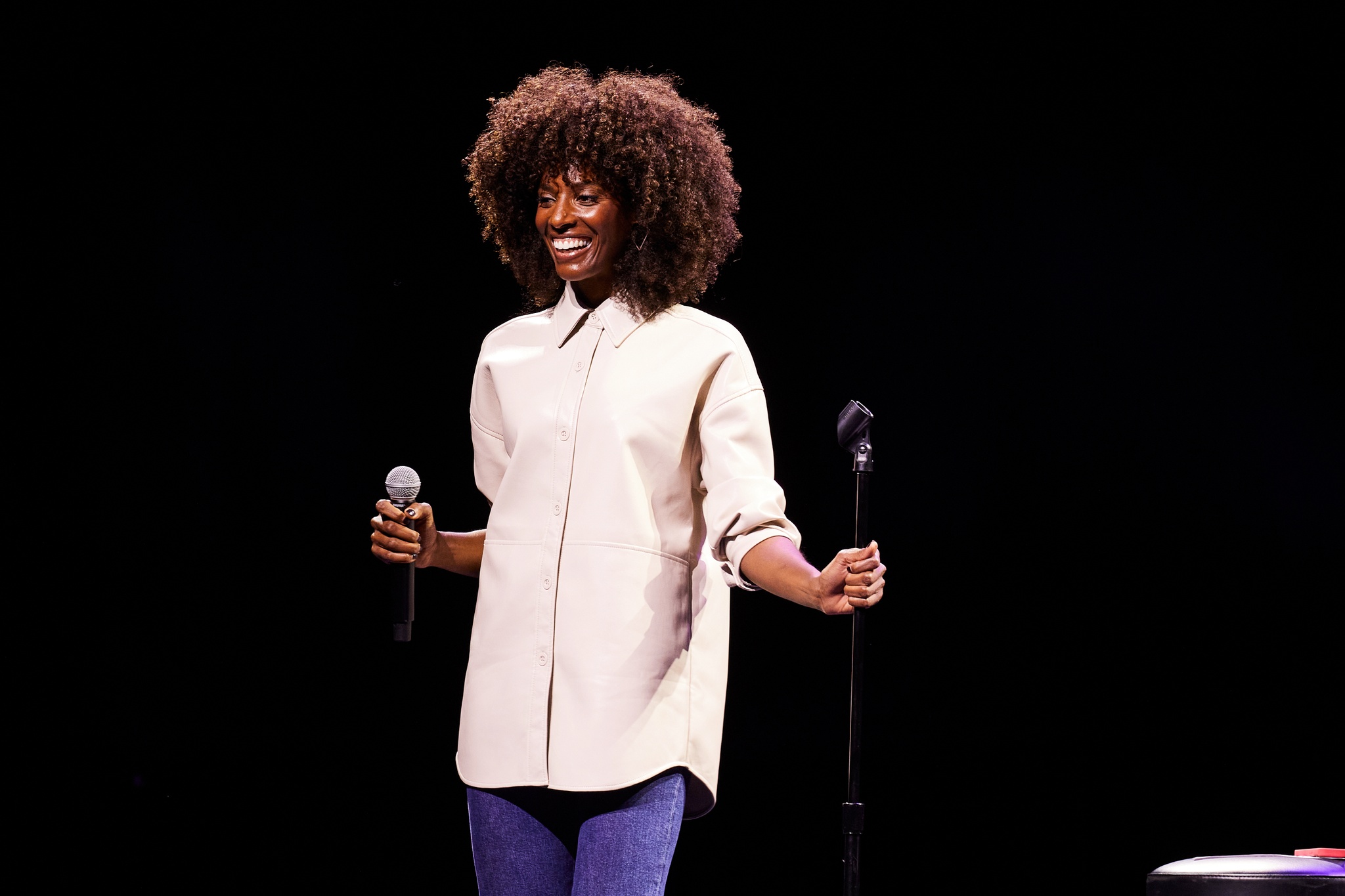A Black woman on stage holding a microphone in her right hand and grasping the microphone stand with her left. She has curly hair styled in a loose Afro and wears a cream-colored button down shirt untucked. 
