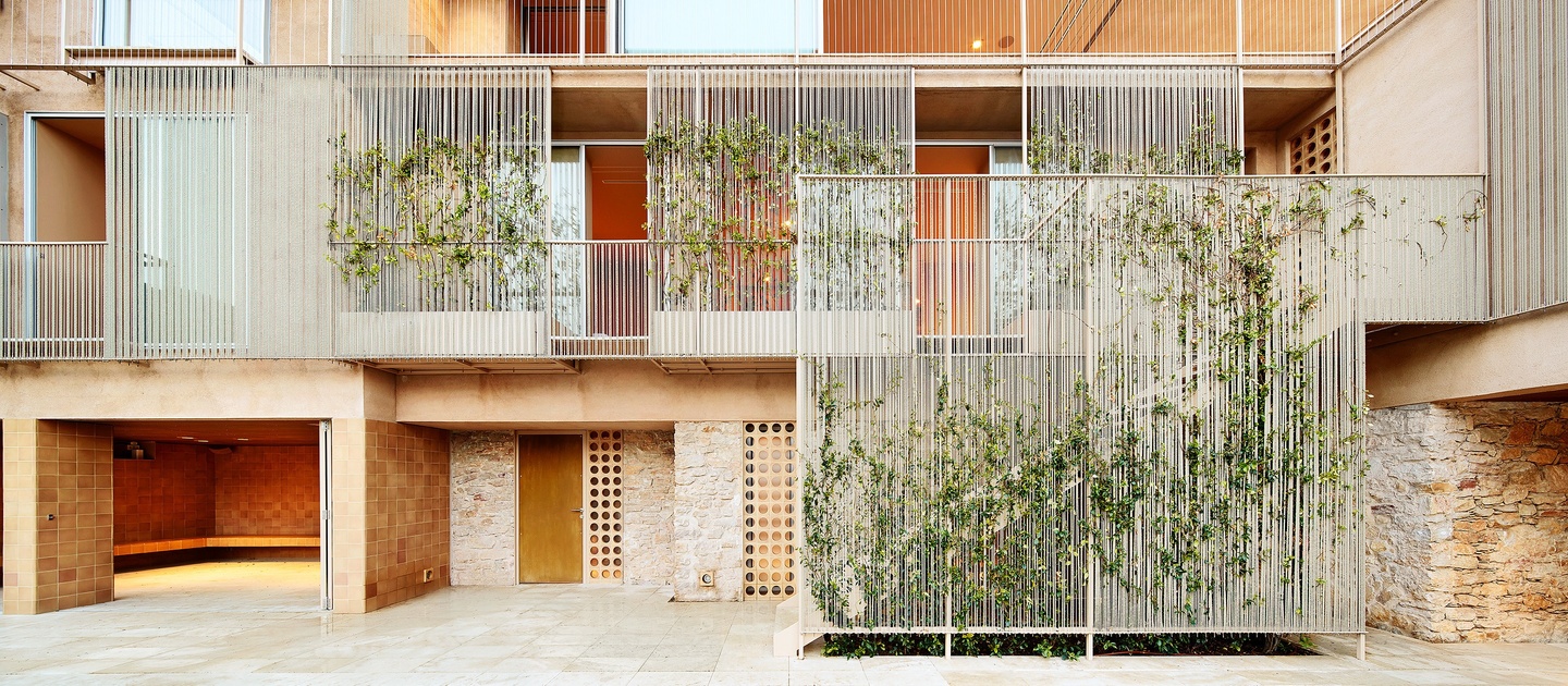 Exterior facade of a multistory story building that's tan with multiple thresholds and some stone work. Multiple screens with small slat openings are in front of sections of the building.