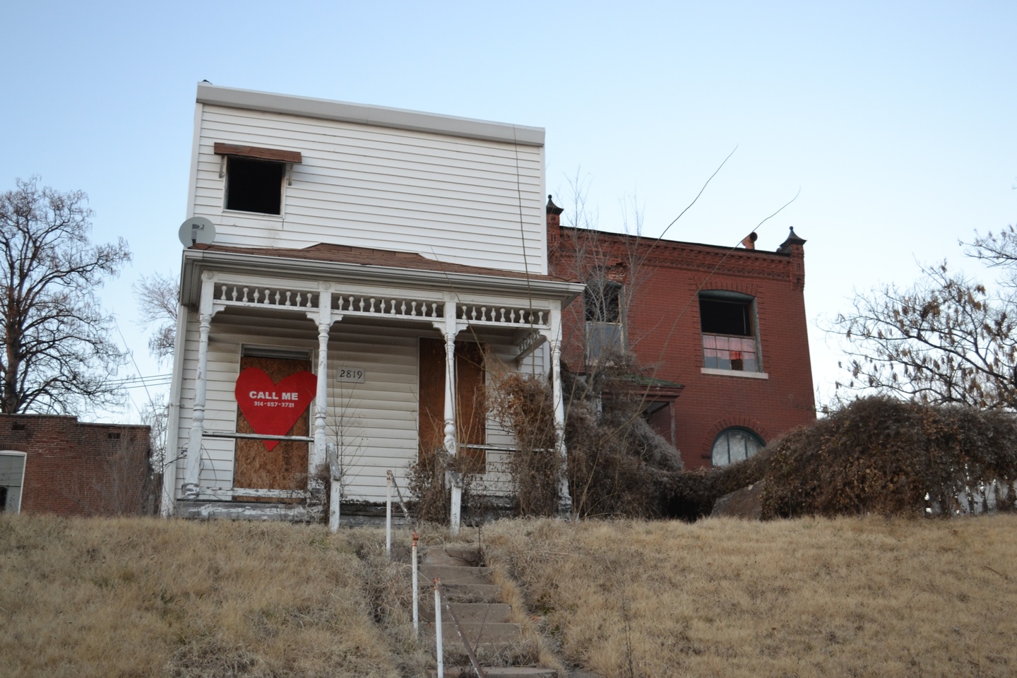 Abandoned house with a large red heart nailed over the boarded up door that reads "Call Me" with a phone number.