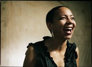 A woman with short hair leans over slightly in a laugh. She wears a frilly tank top blouse and stands against a neutral background.