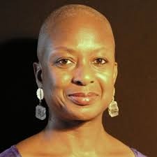 A headshot of Eva Yaa Asantewaa, a Barbadian woman with brown skin who has a shaved head and wears dangling earrings. She smiles warmly without opening her mouth.