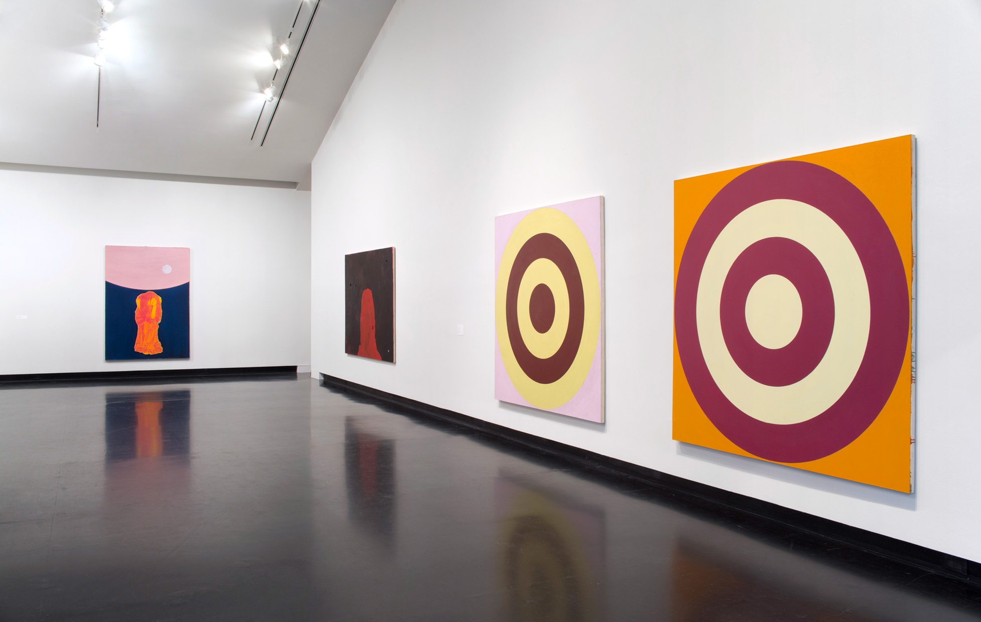 Four large paintings on white walls. Two are abstract paintings of targets and the other two are abstract paintings of a female figure.