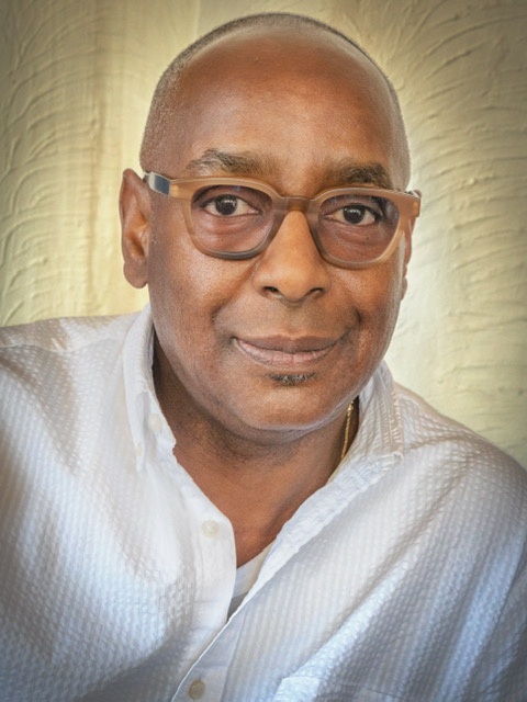 A photo of James King in a white shirt and tan and black framed glasses posing against a cream-colored wall. 
