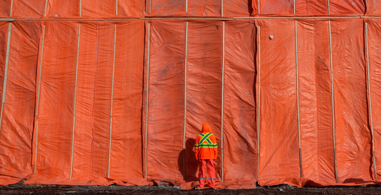 A figure in an orange surveyor's uniform stands with their back to us in front of a large orange tarp