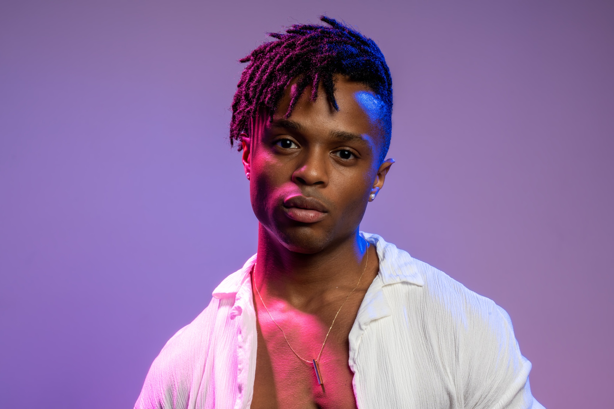Artist John-Michael Lyles looks directly at us in a portrait where we see him from the chest up. He is a black man with short locs falling over his forehead. He wears an unbuttoned white shirt and is cast in dramatic pink and purple light. 