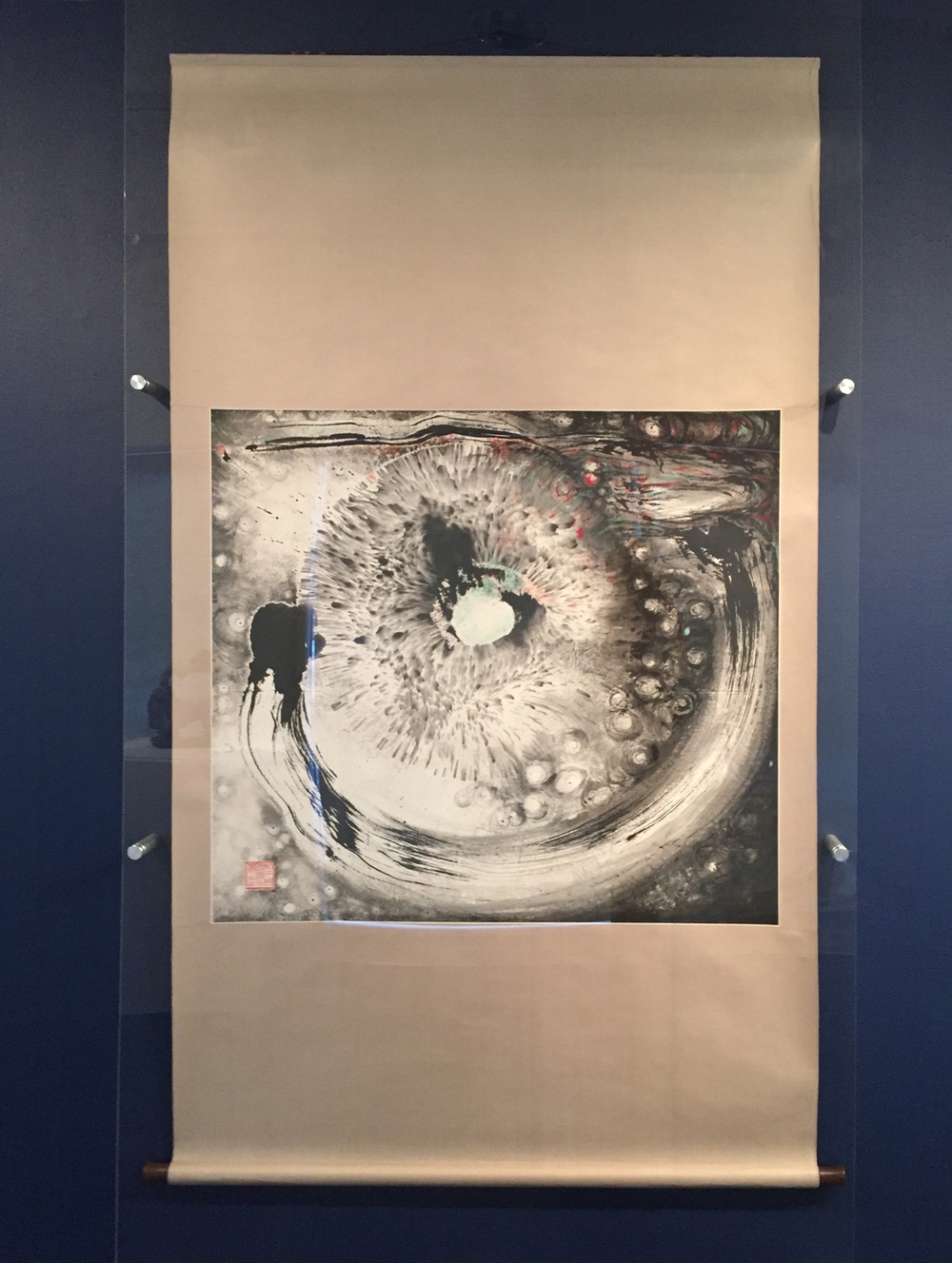 A long tan paper scroll with an abstract, black and white, ink painting in the center of the scroll hung on a dark blue wall.