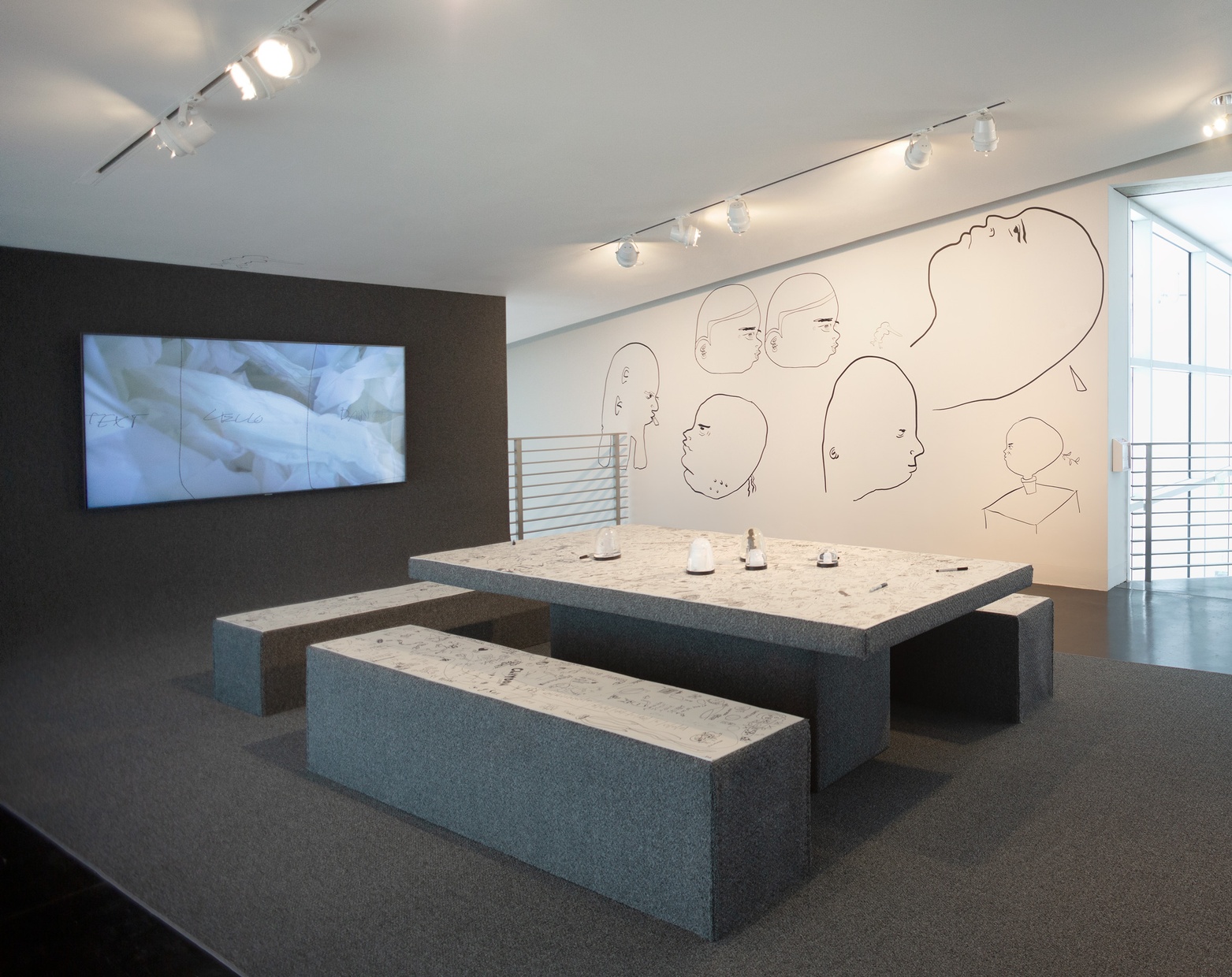 A small gallery features a carpeted wall, seating, and floors, all topped with a white board, one white wall with black line drawings, and a video playing on the carpeted wall.