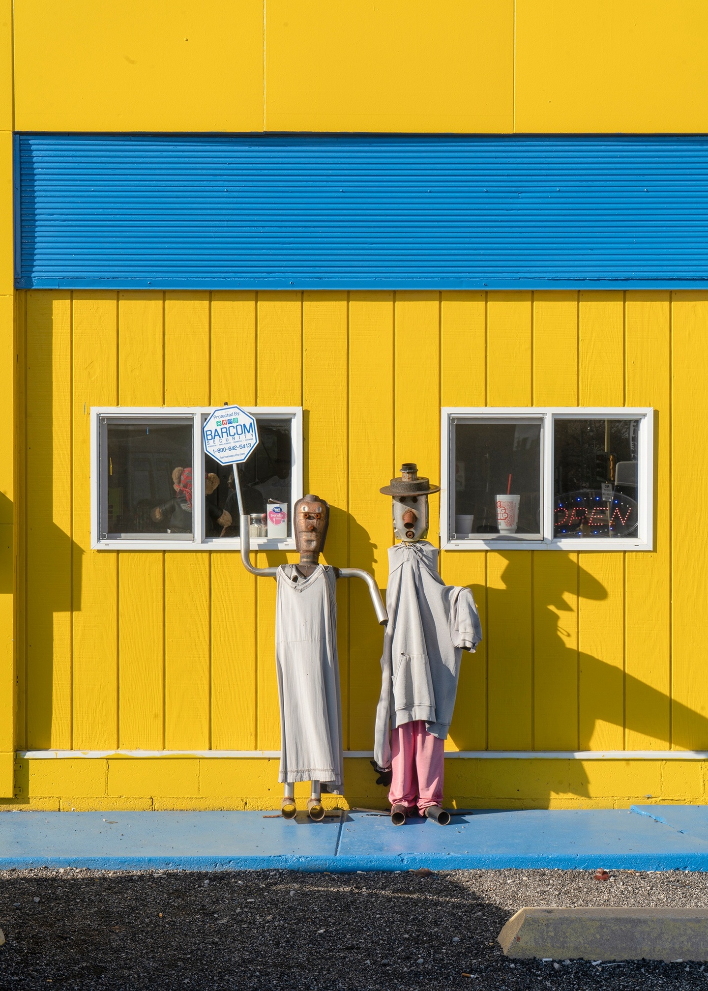 Exterior of a building with wood siding and small windows, painted bright yellow with blue accents. Two humanoid sculptures made of muffler parts dressed like a man and a woman are propped against the wall.