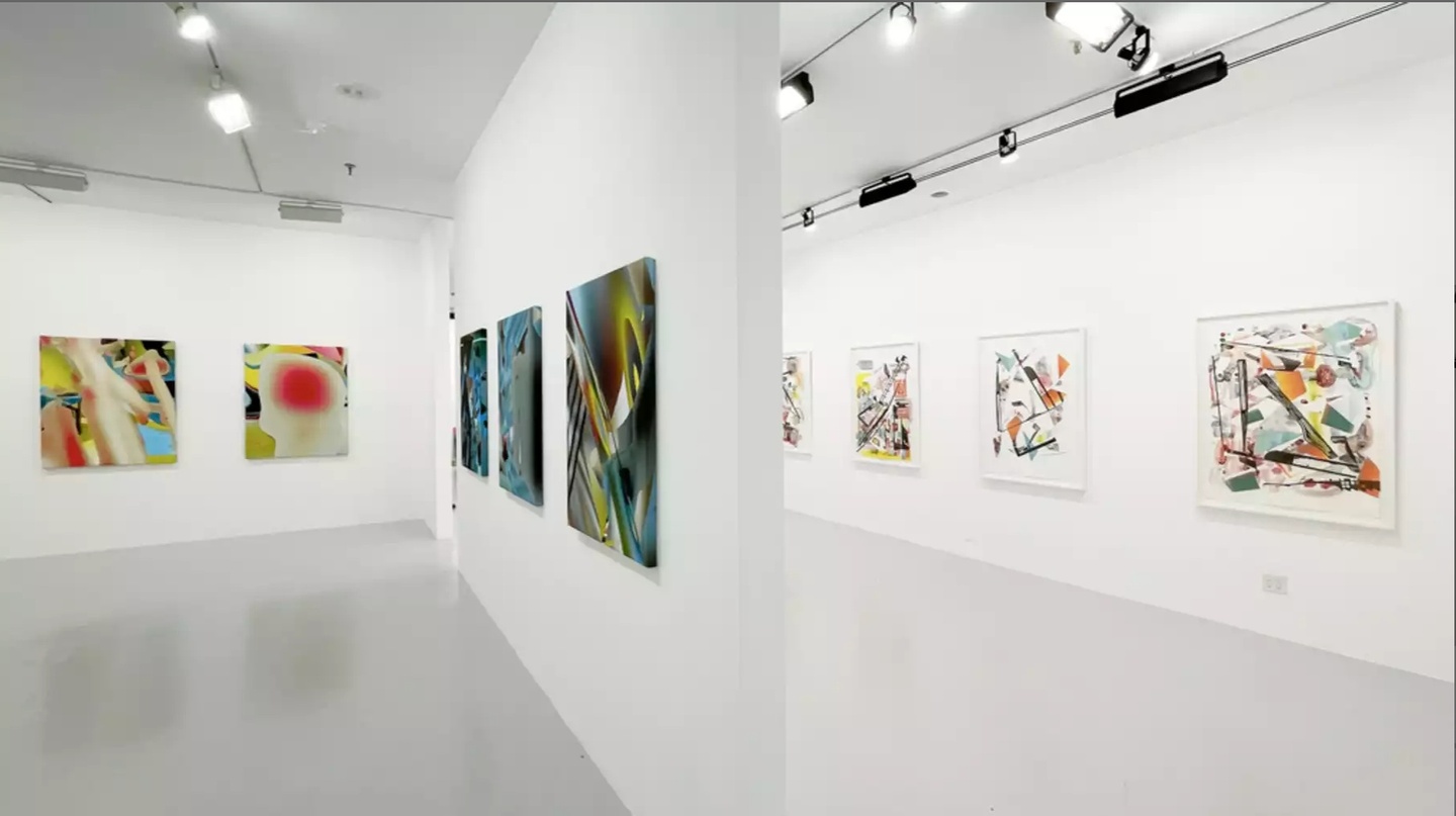 White-box gallery space with a dividing wall; several colorful prints are hung on the walls.