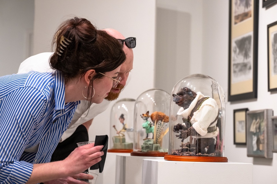 Two gallery attendees peer into a glass vitrine to view a small cartoon bear figurine.