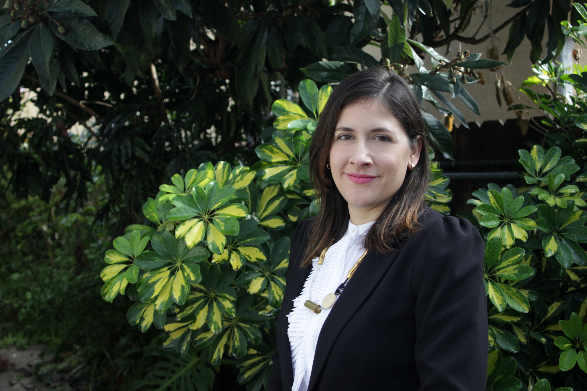 A photo of Marcela Guerrero standing against a background of green leaves. Guerrero wears a black blazer, white blouse, and a long necklace. 