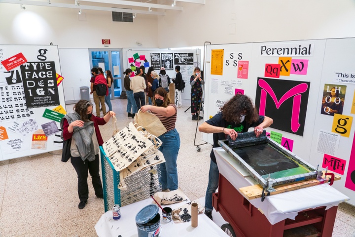 Gallery space with pinboard walls covered in colorful typographic posters and type samples. A pop-up screenprinting station in one corner of the room is printing tote bags.