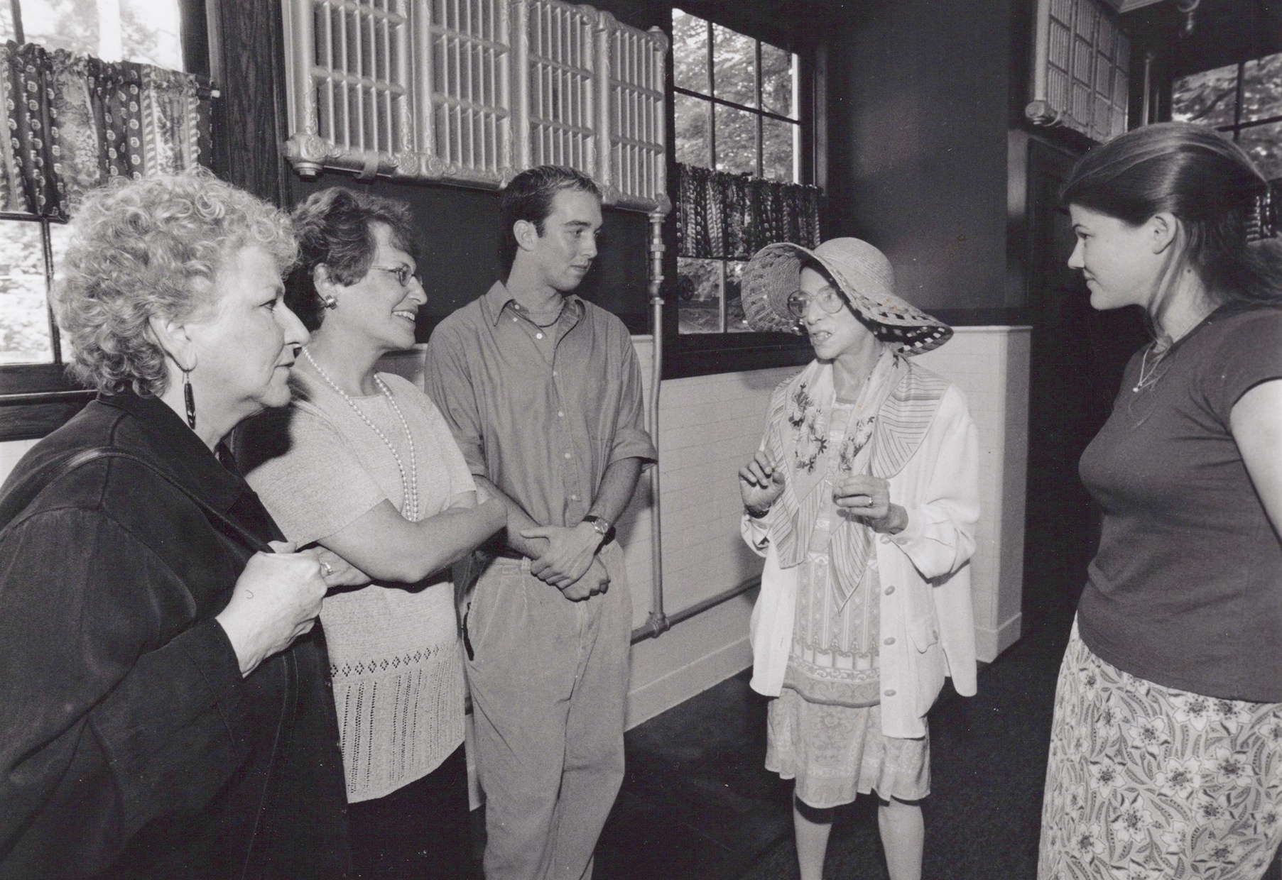 A black and white photograph of an older, light-skinned woman in a large hat talking to a group of people, two older women and two younger people.