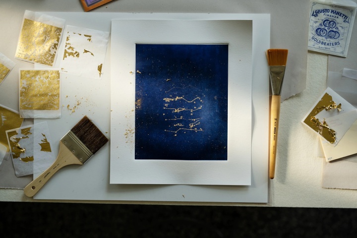 A print with dark blue background and lots of small markings in gold lays on a tabletop framed by two brushes and sheets of gold leaf.