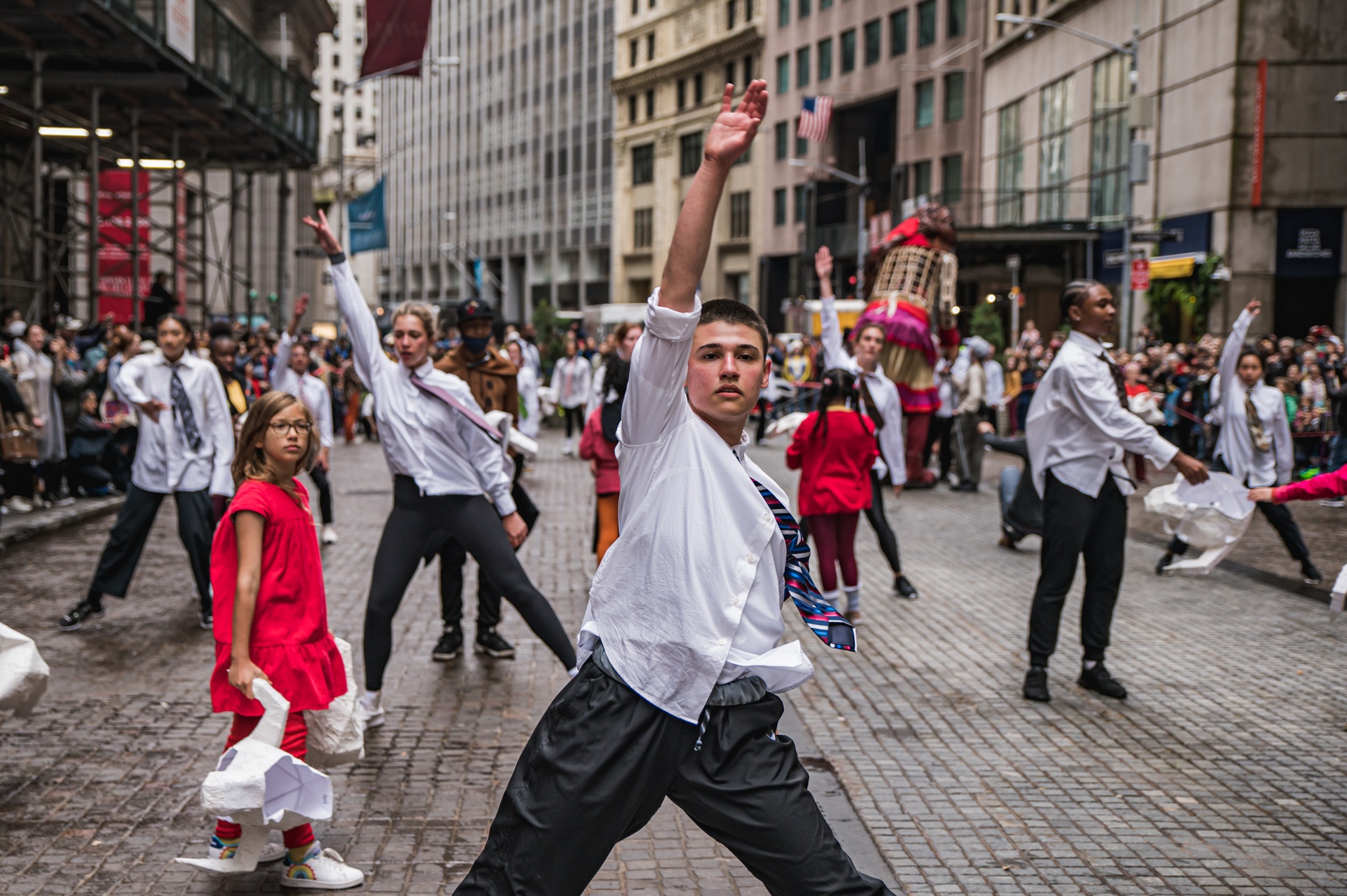 A group of dancers appear on Wall Street, wearing white button ups, black slacks, and ties, with an arm stretched upward. A single dancer with a shaved head can be seen in the foreground. Young children wearing bright colors stand among the dancers and watch on while holding bull head masks. Behind them, the puppet Little Amal can be seen.