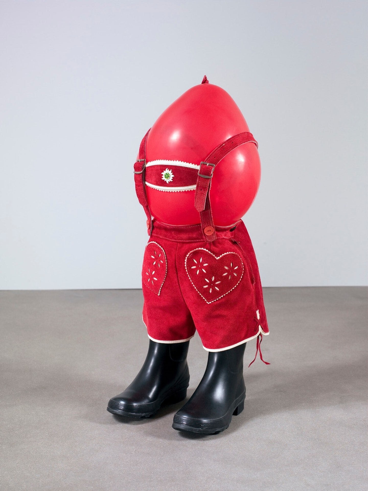 A red inflated balloon fitted with red lederhosen featuring heart-shaped cutouts and a decorative floral motif, secured with suspenders, stands upright in black rubber boots against a plain gray background.