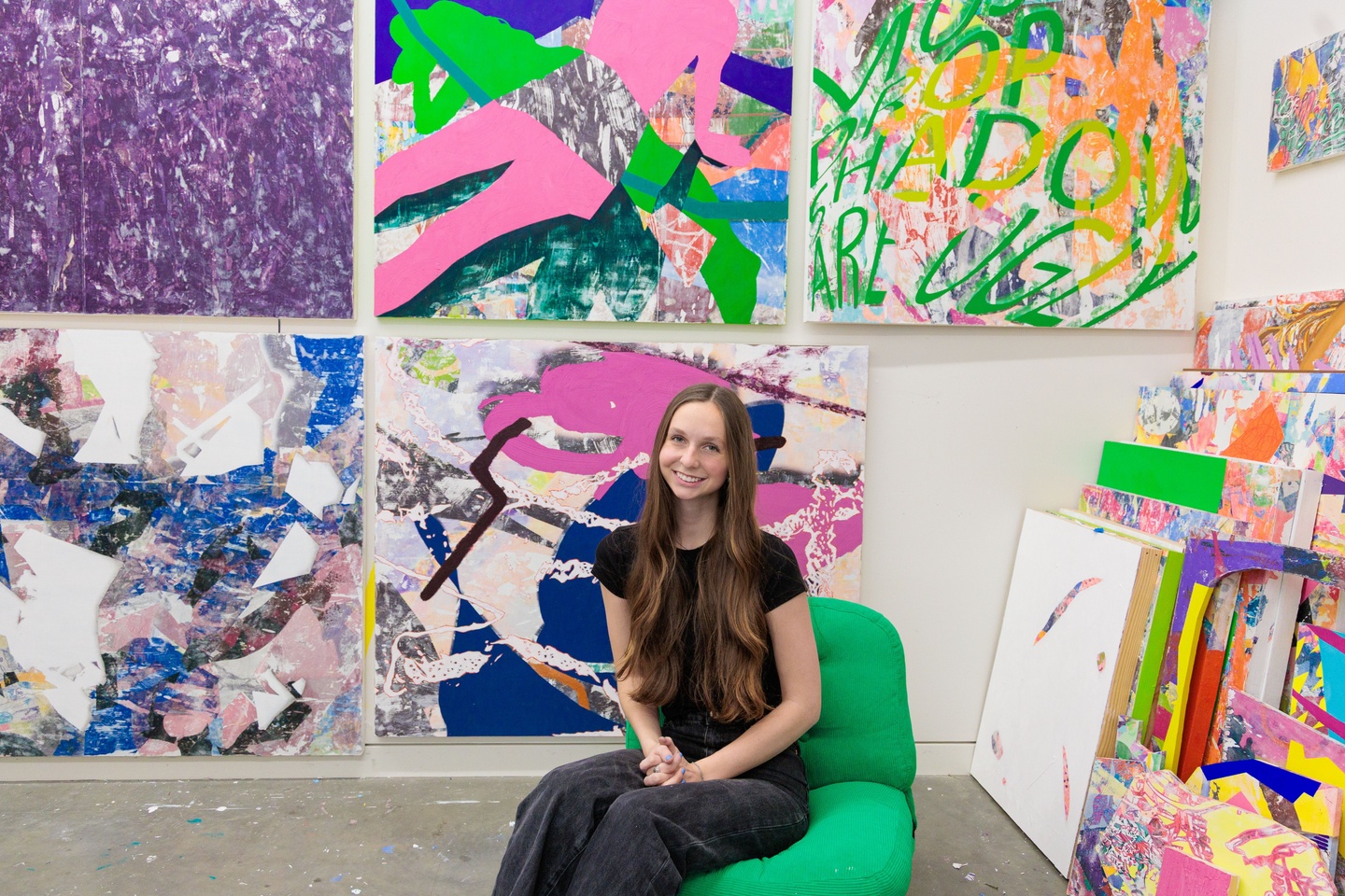 The artist is wearing all black with long hair sitting in a bright green chair. Behind her are several large abstract paintings in bright pinks, purples, greens, blue, yellows, oranges, and more