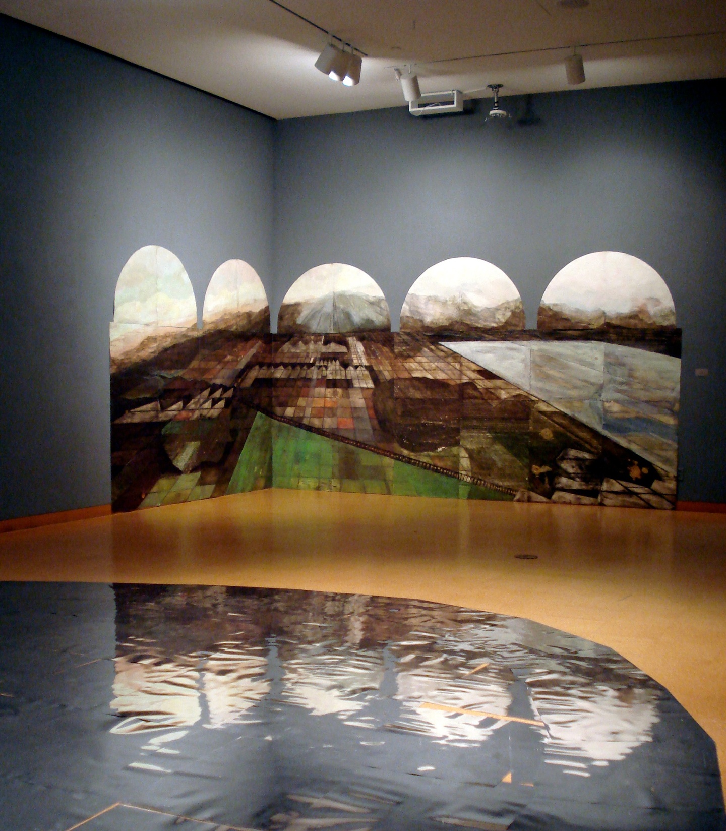 A gallery space: the walls are gray/slate with wooden floors: a landscape is installed along the corner of the space, as if the walls frame the landscape, outlined in five rounded arches; in the foreground on the floor is a reflective surface that shows the reflections of the landscape in the back, as if the glimmering ripples of a pond. Lights on the ceiling illuminate the corner of the space with the landscape, casting a shadow on the ground between the corner and the foreground.