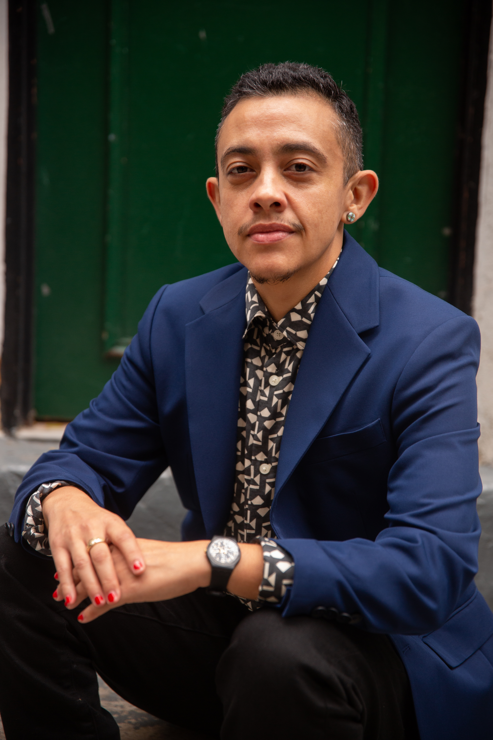 A portrait of J. Soto who wears a blue blazer and poses sitting with arms propped on his knees. He wears watch on one wrist and pink nail polish. He looks directly at us. 
