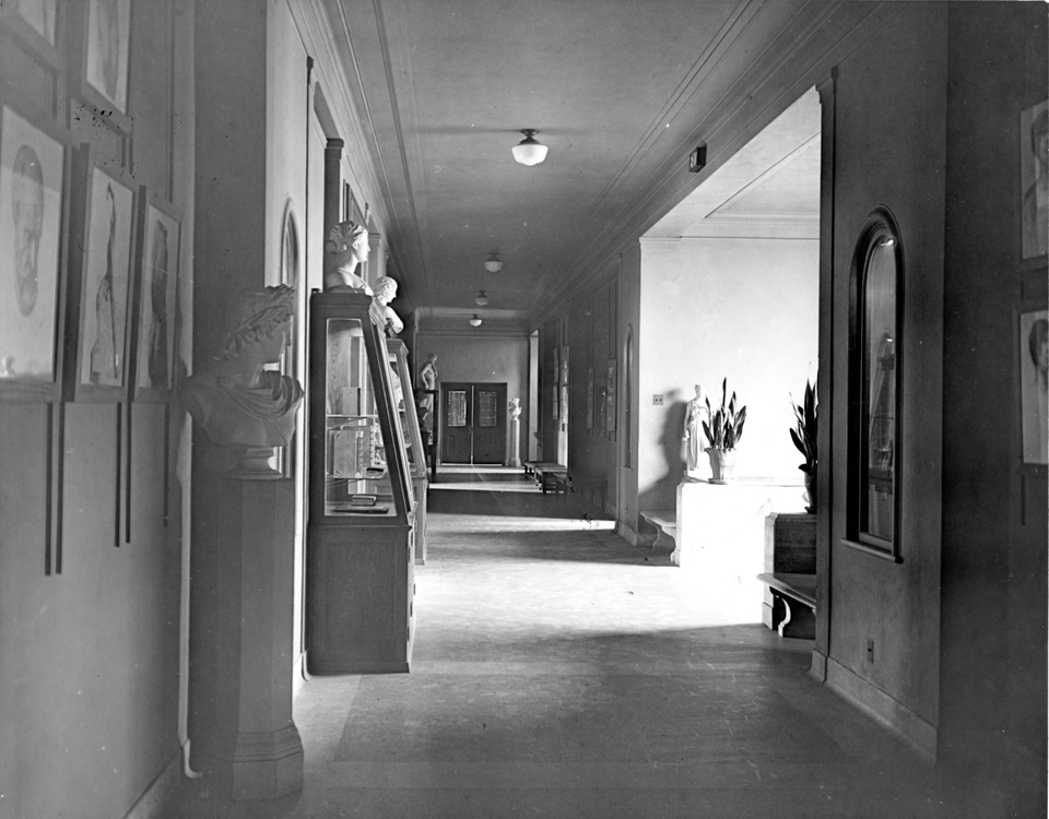 Black and white photo of a hallway filled with sunlight, displaying various paintings and classical sculptures.