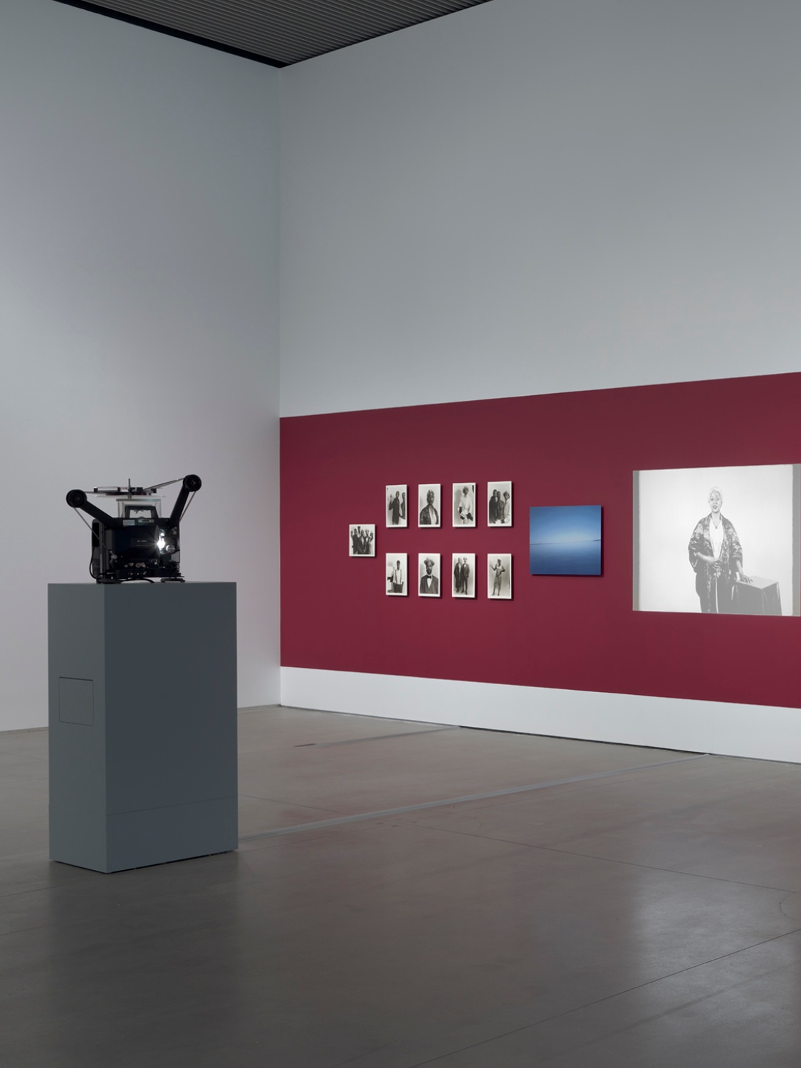 A film projector sits on a plinth at a distance from a red gallery wall with photos of different sizes hung on it