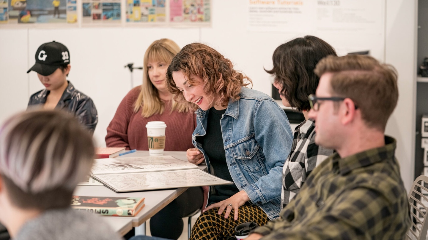 A group of graduate students and faculty sit around a conference table in an illustration studio, engaging in lively discussion.
