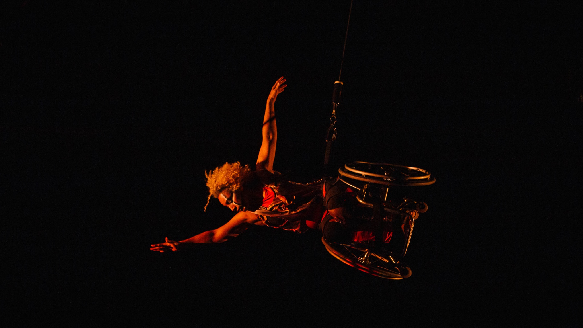 Alice Sheppard flies in her wheelchair, body parallel to the stage as she reaches one arm high and extends the other to the side. She dances in a glowing red shadow. She is a multiracial Black woman with coffee colored skin and short curly hair; she wears a red and gold bodysuit, black and white lines adorn her face.
