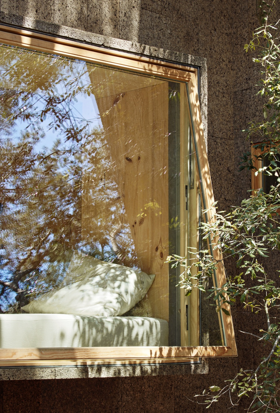 Closeup of the large window of the cork house. A white bed with white pillows are seen inside the house by the window 