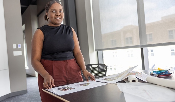 Individual stands smiling across the table with architectural drawings at their fingertips.