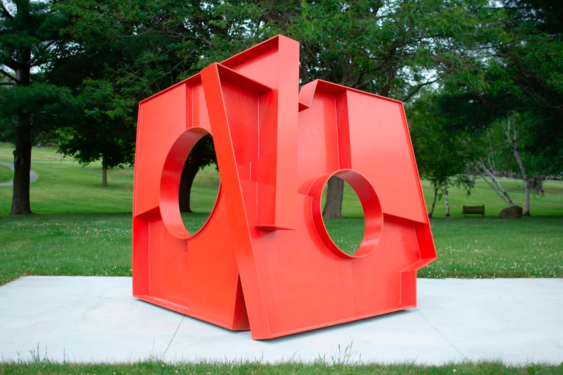 A large, red sculpture of two rectangular shapes conjoined at an angle with large holes in the middle of each plane sits on a concrete slab in a grassy field with trees.