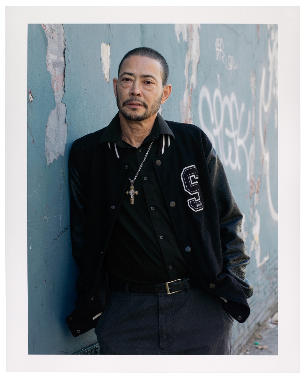 A photograph of an older, Asian person with short buzz cut hair in a black letterman jacket wearing a metal cross leaning against a blue, cement wall. 