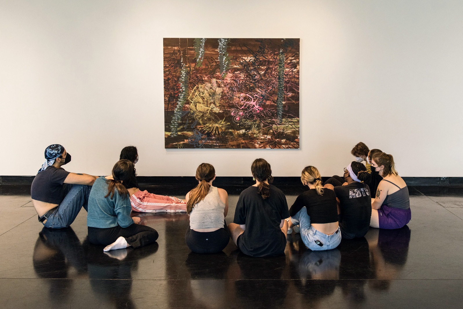 A group of college students, with their back to the camera, sit on the floor looking up at an abstract painting hung on a white wall.