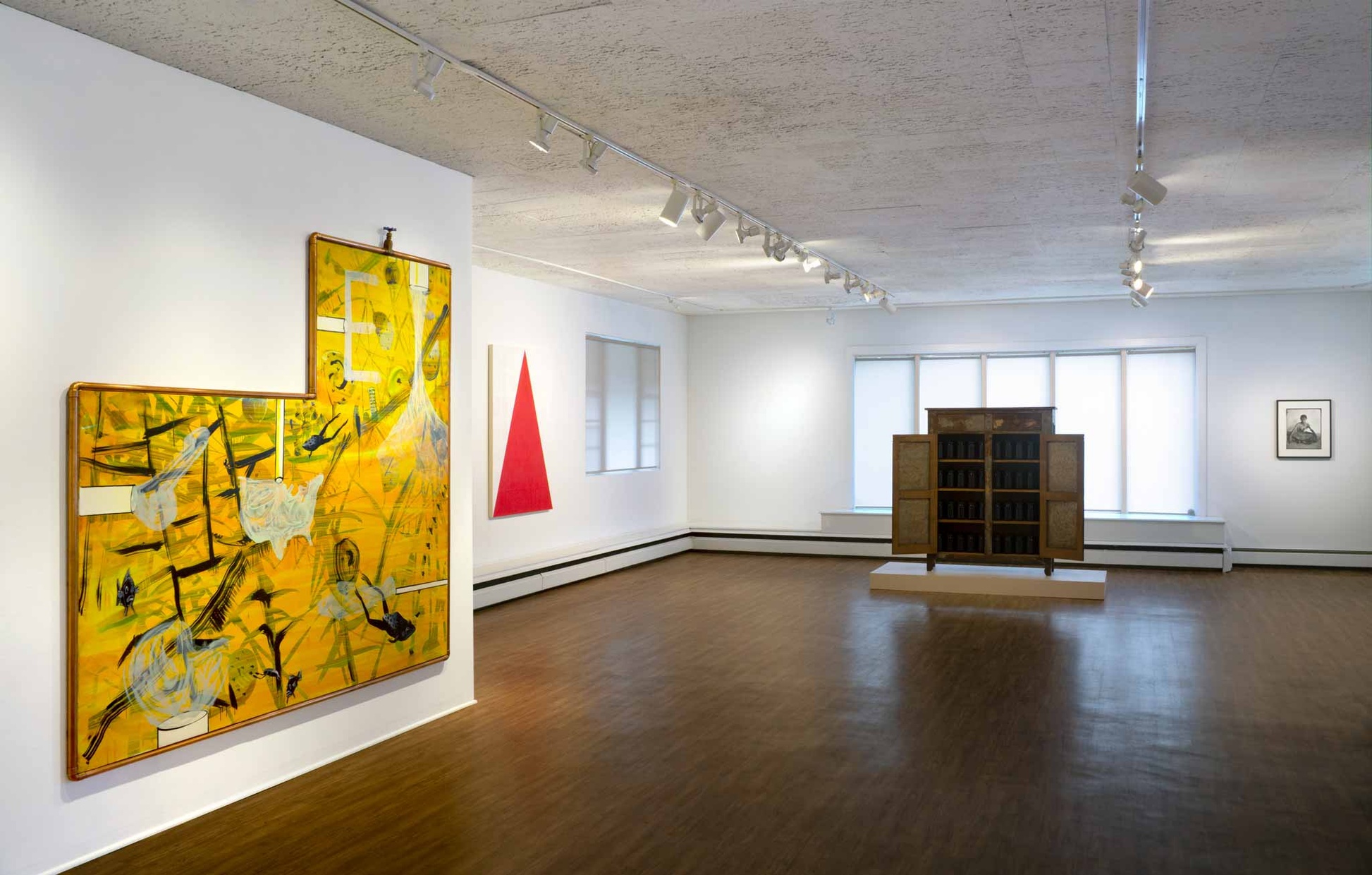 A large room with large paintings hanging on white walls and a cabinet on display near the back wall.