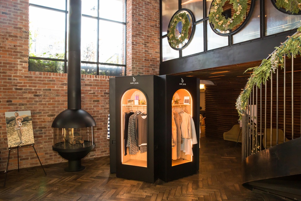 Pop-up space, showing two faces of the structure, which is framed in black with arched, backlit windows showing clothing inside. The space is set up inside a red-brick building, to the left of a skinny fireplace and larged, paned window.