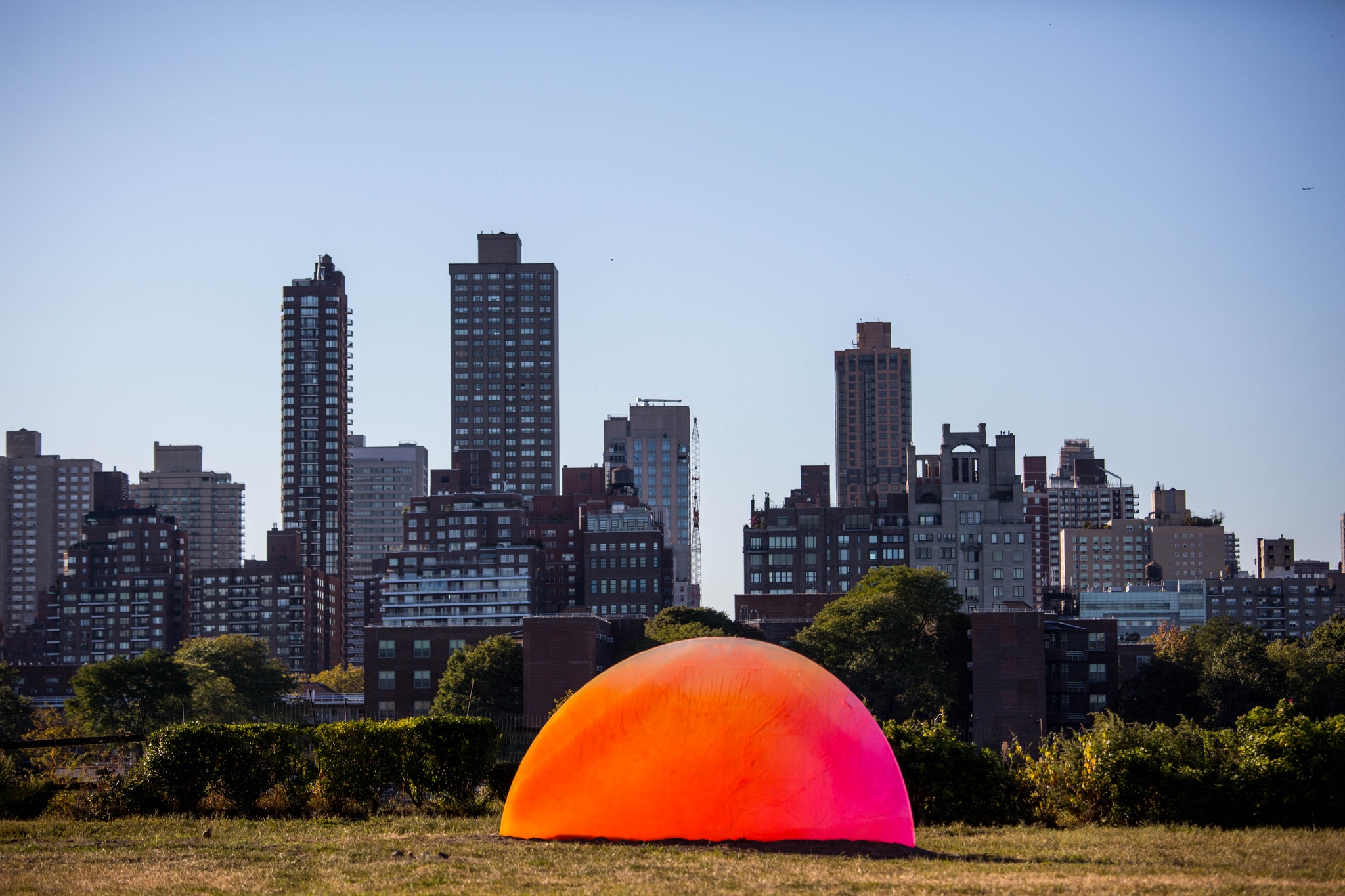 A large half-circle panel sits in a field with a city skyline in the background. The half-circle is colored with a gradient of fluorescent oranges that fades into pinks and purple, evoking a sun rising out of the earth.