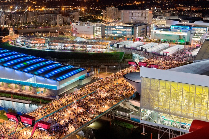 Nightime aerial photo of the scene at the London Olympic and Paralympic Games. The scene includes a venue with bight blue rooftop lights to the left, and a multilevel rectangular stucture with tall, yellow windows on the second floor. Between the two buildings is an open spaces dotted with large crowds of people. In the background is a view of more buildings lit up.