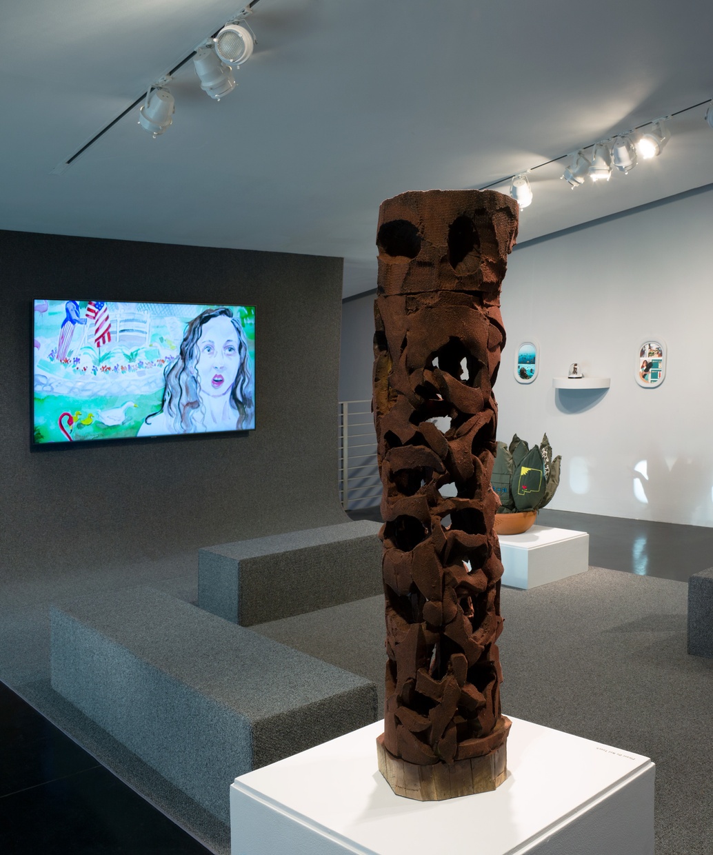 A vertically oriented sculpture sits on a plinth, and in the background is a television showing an image from a watercolor animation, a sculpture of a cactus, and framed collages.