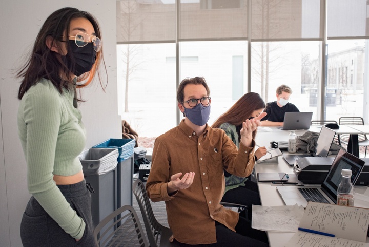 Seated instructor speaks to a standing student and gestures to something off camera. The table in front of them is covered with laptops, scanners, and sheets of tracing paper with drawings.