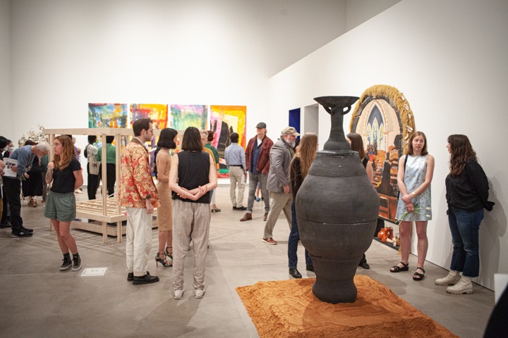 Gallery space displaying a large black urn, a painting built into a frame with shelves and grass fringe, a rectangular wooden framework, and a set of four colorful abstract paintings.