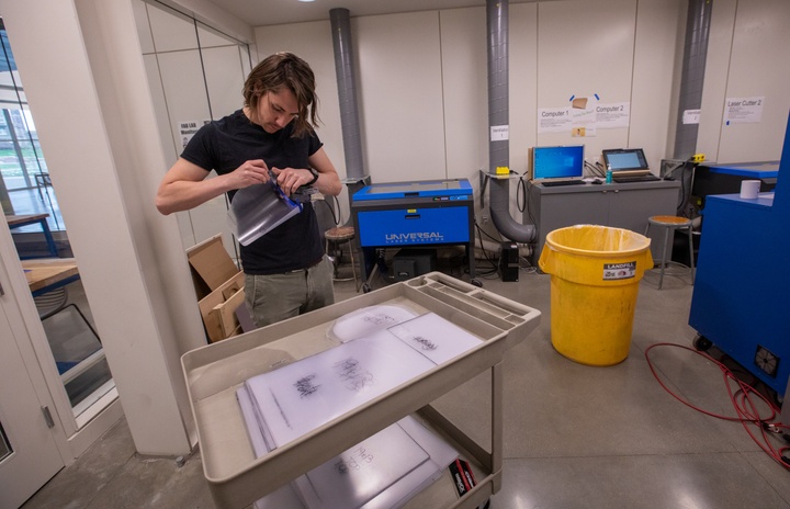 Person assembles a plastic face shield at a cart with stacks of pre-cut face shield pieces.