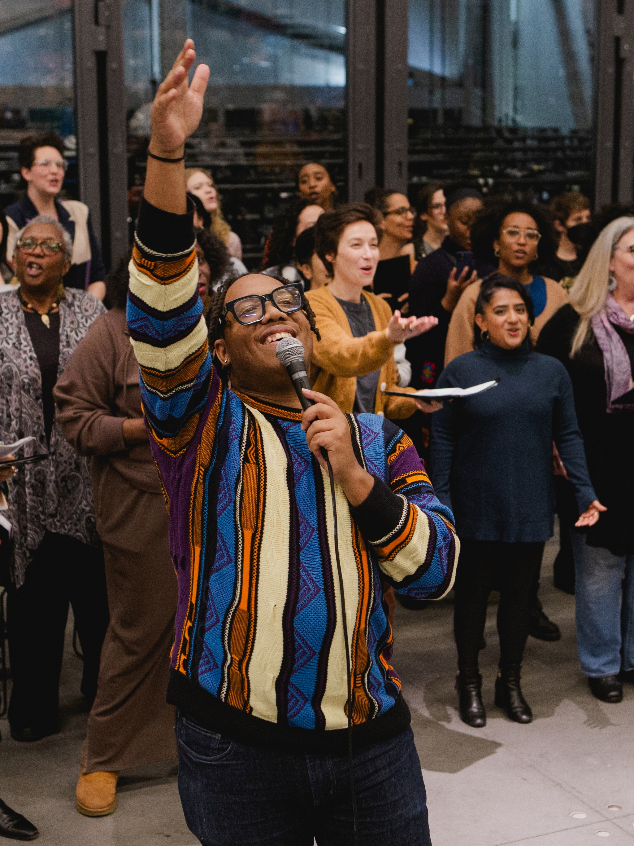 The Fire Ensemble choir rehearses in an event space at The Shed. In front of the choir who is assembled standing in rows, the choir's artistic director Troy Anthony stands holding a microphone reaching his other hand high above his head with a broad smile on his face. 