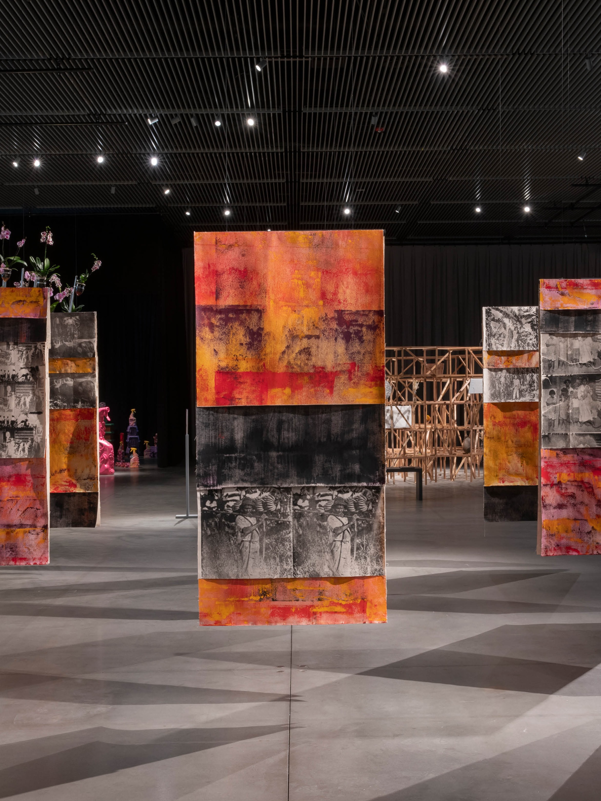 Nine painted fabric panels hang from a gallery ceiling in a three by three grid. The panels are painted in orange and black bands, with screen printed sections with black and white images of banana plantation workers in the 1920s. 