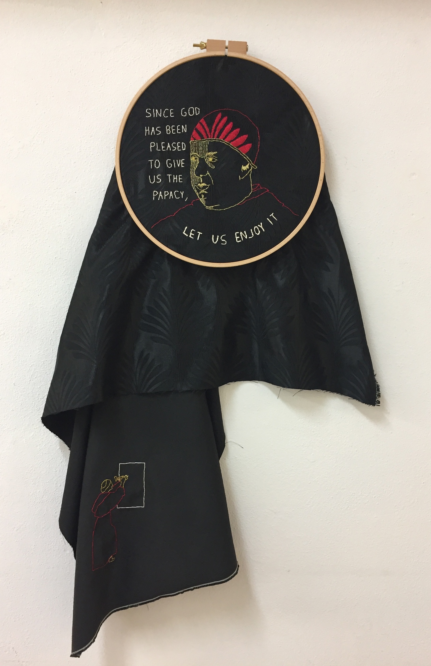 Piece of black cloth in an embroidery hoop, with an embroidered image of the pope's head in the center. Text around it reads "Since God has been pleased to give us the papacy, let us enjoy it." The bottom corner of the fabric has an embroidered image of a monk beginning to engrave a tablet.