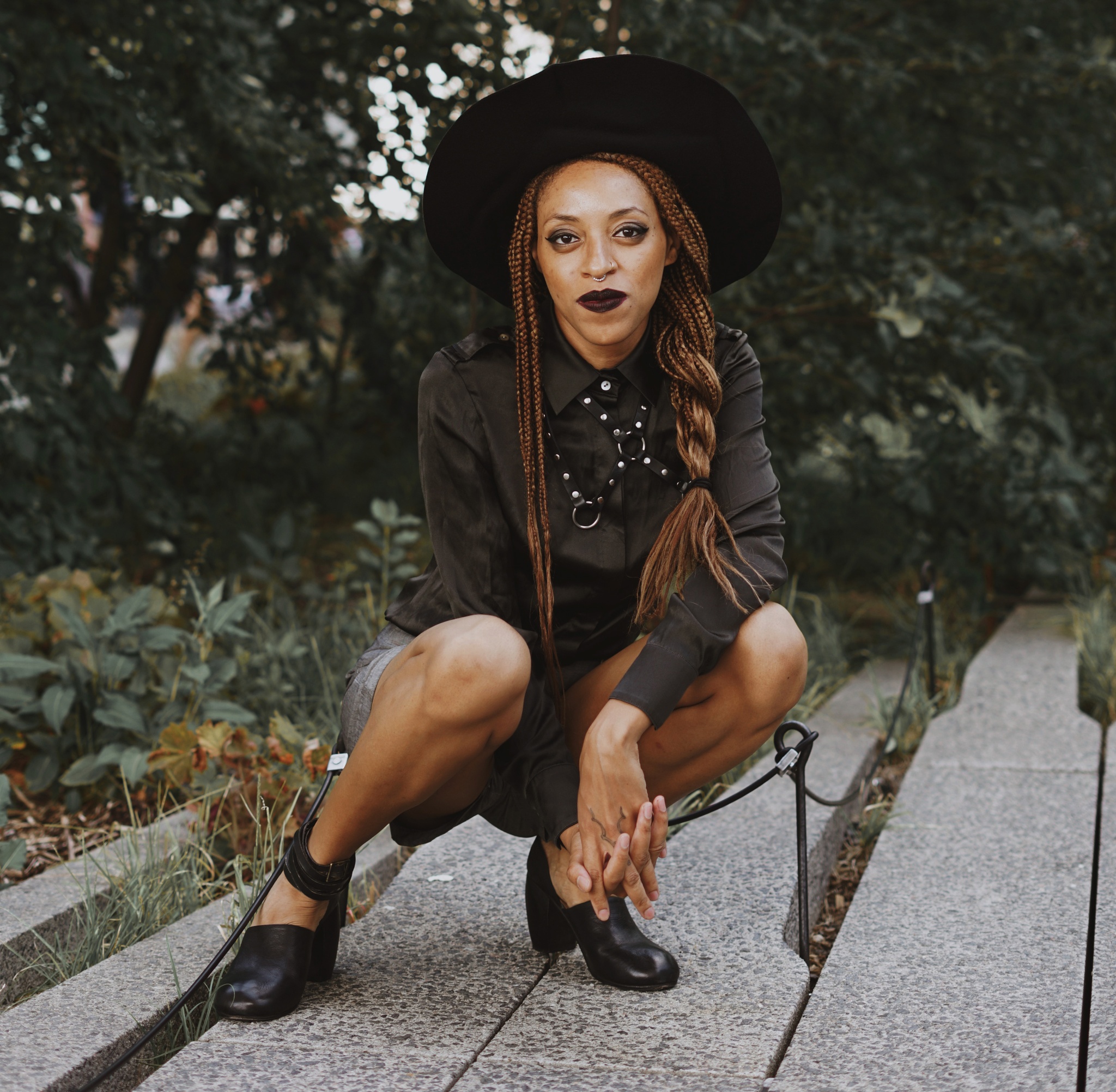 A photo of the artist Ayanna Dozier crouching on a concrete path in front of green trees, grass, and bushes. Dozier wears a wide-brimmed black hat over her long braided hair.