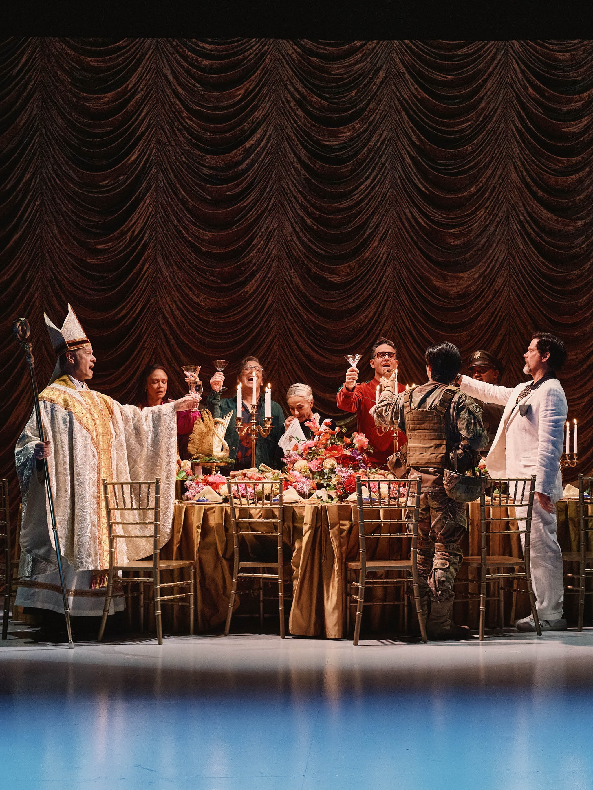 The cast of a play dressed as members of a dinner party lift glasses in a toast as they stand around a long table. At one end stands a guest dressed in white Catholic clerical robes and at the other end a man in a white suit stretches his arm out.