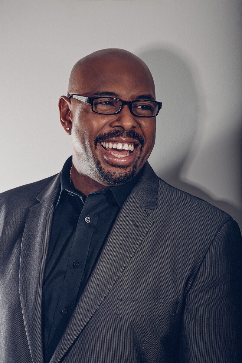 Musician Christian McBride stands smiling against a gray wall where his shadow is cast. He has a bald head and wears a dark gray suit, a dark shirt, and glasses. 