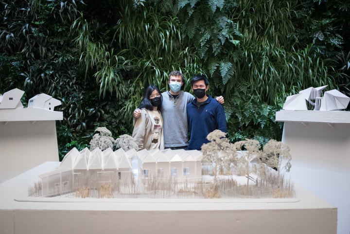 Three people smile and pose behind a table containing an architectural model of a low building.