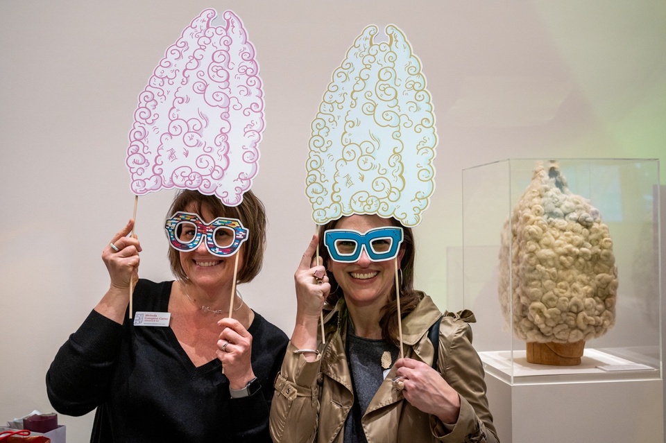 Two people hold up 2D props of a tall, curly wig and wild eyeglasses. Behind them in a vitrine is the wig itself.