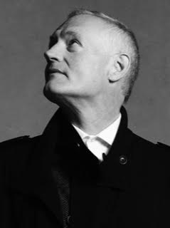 A black and white portrait of a white man with short hair buzzed even shorter on the sides. He looks up and away out of the frame of the image and wears a dark jacket buttoned to the top where a white collared shirt peeks out. 