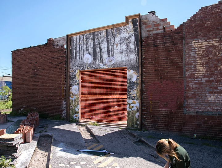 Outdoor brick wall flyposted with a blackwhite image of a forest with dandelions. Entrance into the brick building is covered with orange construction fence